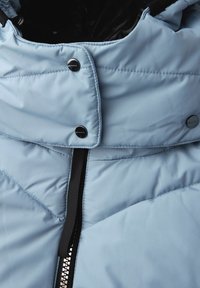 Giacca puffer azzurro chiaro con colletto alto, chiusura frontale con zip, bottoni a pressione e texture trapuntata con dettagli neri.