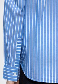 Cămașă cu dungi bleu deschis din bumbac, cu dungi verticale albe, mansetă cu nasturi și tiv rotunjit cu textură subtilă.