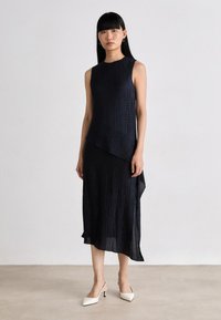 Woman with straight black hair wearing a sleeveless textured black midi dress and white pointed-toe slingback heels standing on a light floor.