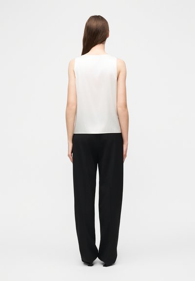 Γυναίκα με μακριά καστανά μαλλιά, ντυμένη με λευκό αμάνικο τοπ και φαρδιά μαύρη παντελόνια, στέκεται με την πλάτη προς το μέρος μας μπροστά σε ένα απλό φόντο.