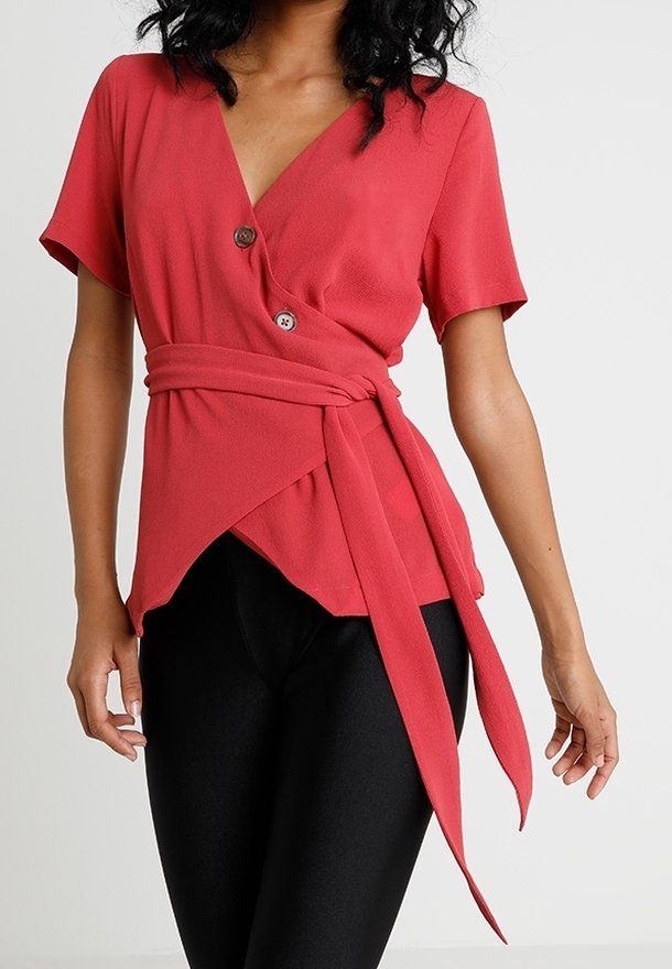 Haut cache-cœur rouge à manches courtes, avec deux boutons à la taille et un détail de nœud. Le tissu a une texture lisse. Associé à des leggings noirs.