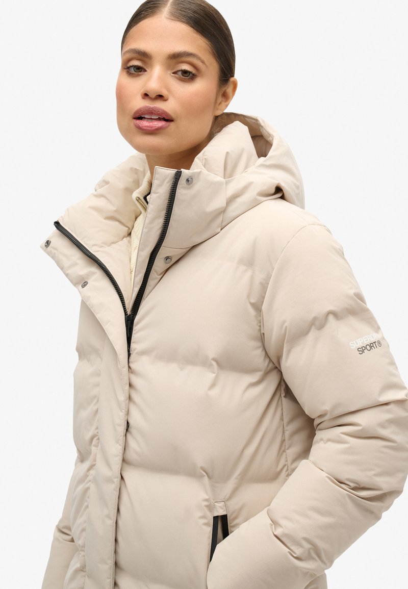 Jasno beżowa kurtka puchowa z wysokim kołnierzem i kapturem, wyposażona w zamek błyskawiczny z przodu oraz boczne kieszenie, z czarnymi detalami zamka.