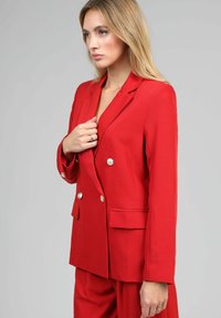 Femme portant un blazer rouge à double boutonnage avec des boutons dorés, ajustant son revers, se tenant devant un fond gris clair uni.