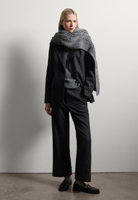Zwarte getailleerde blazer en broek, grijze gebreide trui en een gestructureerde grijze sjaal. Zwarte instappers maken de monotoon geklede outfit compleet.
