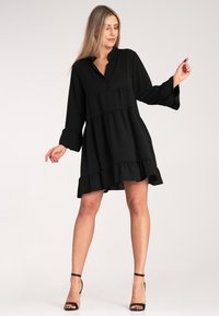 Czarna sukienka z kołnierzykiem, długimi rękawami i falbaną u dołu. Wykonana z miękkiego materiału, posiada warstwowy krój i luźny fason.