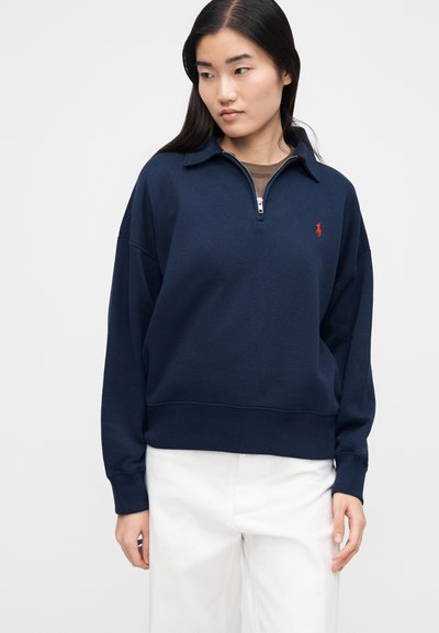 Γυναίκα που φοράει μπλε σκούρο πουλόβερ με φερμουάρ στο ένα τέταρτο και κόκκινο λογότυπο στο στήθος, μαζί με άσπρο παντελόνι, στέκεται μπροστά σε απλό φόντο.