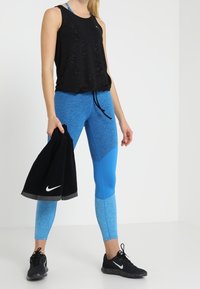 Czarny sportowy top bez rękawów z fakturowanym wzorem, w połączeniu z niebiesko-turkusowymi legginsami z kolorowym blokiem; trzyma czarny ręcznik.