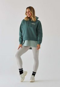 Ciemnozielona bluza z kapturem i sznurkiem, nałożona na jaśniejszą zieloną bluzkę, w połączeniu z białymi legginsami i kremowymi sneakersami. Widoczne czarne skarpetki w paski.