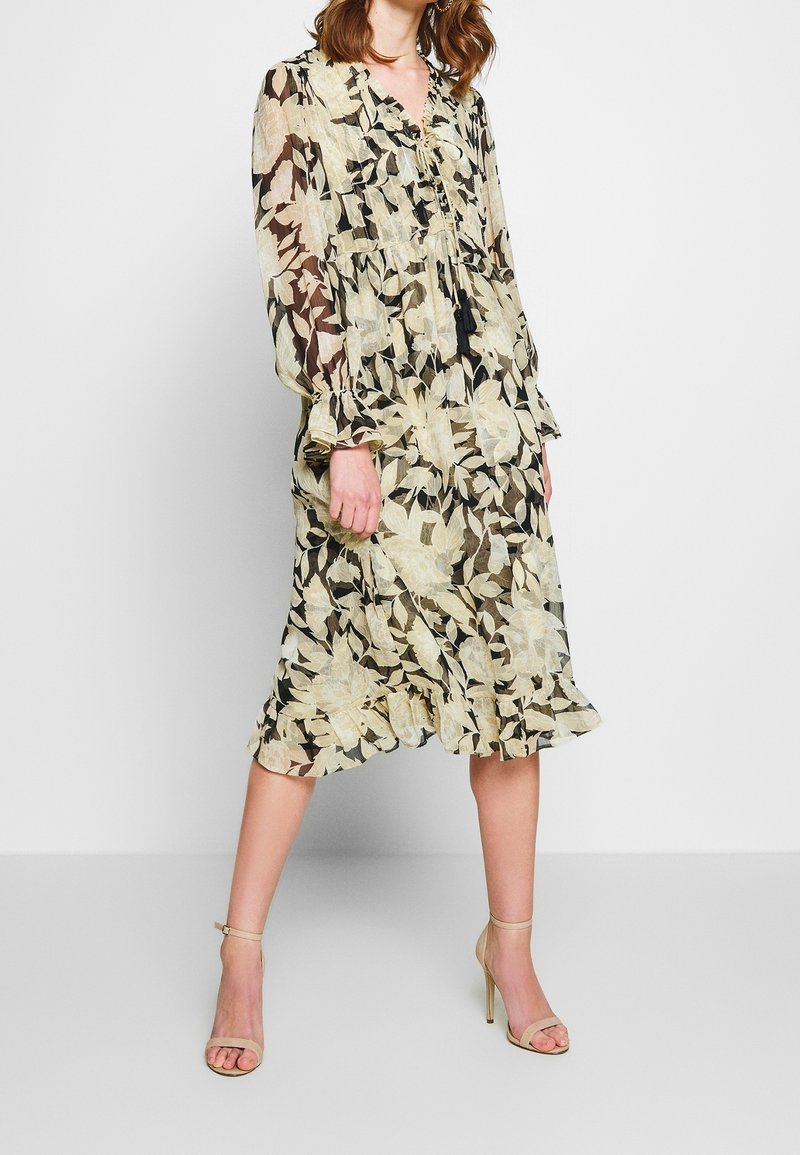 Robe longue à manches longues, transparente et fleurie, en crème et noir, avec une taille froncée et un ourlet volanté, complétée par un lien à pampilles et une texture subtile.