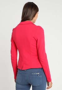 Veste rose ajustée avec un col encadré, manches longues, design épuré et texture lisse. Le dos présente une couture verticale et une légère coupe cintrée.
