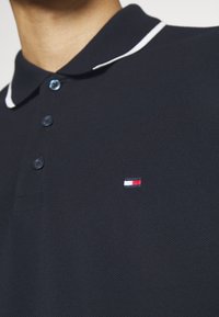 Polo shirt bleu marine avec col blanc, patte de boutonnage à trois boutons et tissu texturé. Présente un petit logo brodé en rouge, blanc et bleu sur la poitrine.