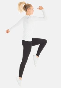 Weißes langärmliges Sportshirt mit strukturierten Ärmeln, kombiniert mit schwarzen Leggings und weißen Sportschuhen; dynamische Pose, die Bewegung zeigt.