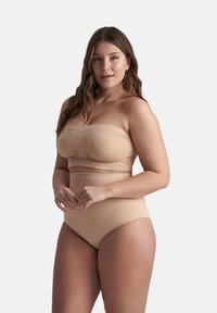 Donna con capelli lunghi marroni che indossa shapewear beige senza spalline, in piedi con le mani delicatamente intrecciate contro uno sfondo chiaro semplice.