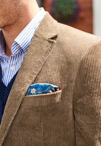 Homme portant un blazer en velours côtelé marron, une chemise rayée bleue et blanche, un pull bleu marine et un mouchoir à fleurs dans la poche du blazer.