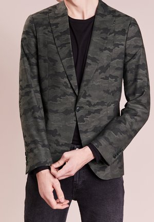 Man draagt donker groen en zwart camouflage patroon blazer over zwart overhemd, mouw vooraan met handen aanpassend.