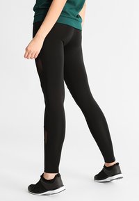 Leggings atléticos negros con paneles de malla laterales. Hechos de un material elástico, con un diseño ajustado y una cintura alta. Combinados con zapatillas negras.