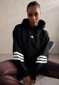 Sweatshirt preto com realces brancos em três riscas nas mangas. Possui um bolso frontal e um logótipo minimalista no peito. Textura suave.