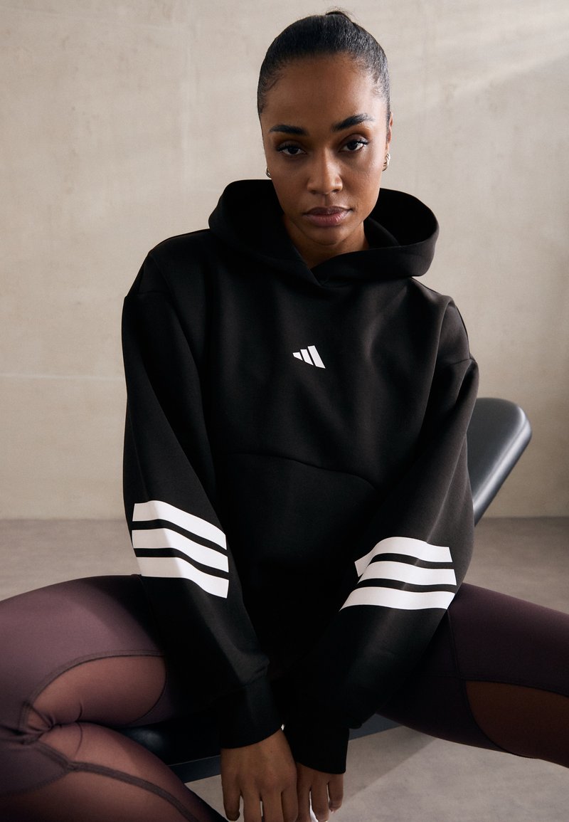 Sweatshirt preto com realces brancos em três riscas nas mangas. Possui um bolso frontal e um logótipo minimalista no peito. Textura suave.