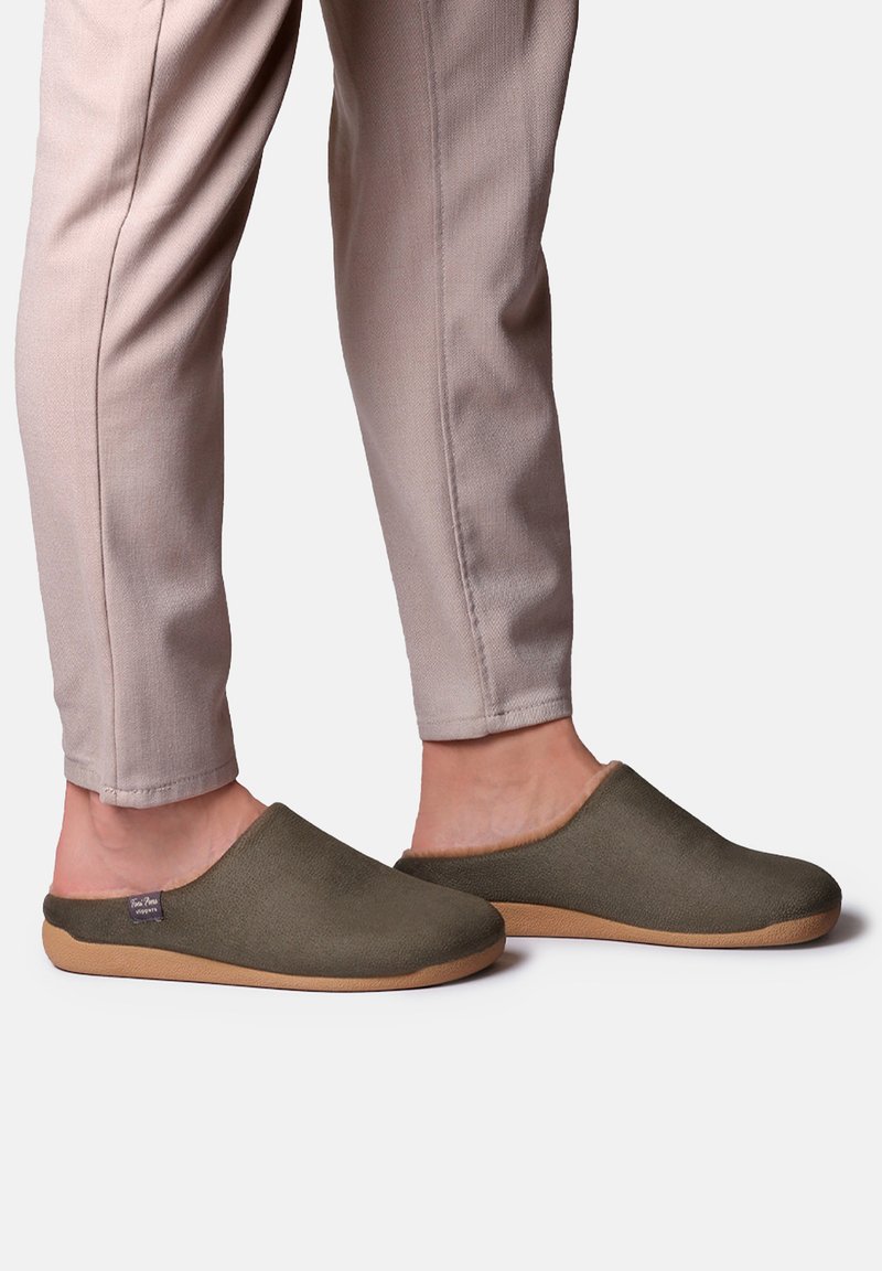 Personne portant un pantalon beige et des chaussons verts à enfiler avec des semelles marron, debout sur un fond blanc.