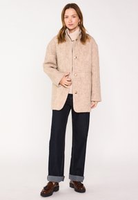Giacca in misto lana beige con taglio dritto, grandi bottoni e tasche applicate, abbinata a jeans denim scuri e scarpe marroni.