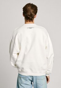 Sudadera crema de corte relajado, con hombros caídos y puños acanalados. Presenta un logo impreso en la parte delantera y trasera. Combinada con jeans azul claro.