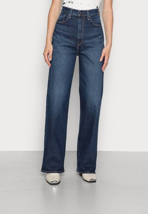 Personne portant un jean droit taille haute bleu foncé et des chaussures blanches avec des bouts métalliques pointus, debout sur un sol gris.
