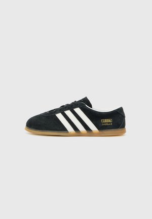 Czarny sneaker Adidas Gazelle z białymi trzema paskami i gumową podeszwą, widok z boku na jasnoszarym tle.