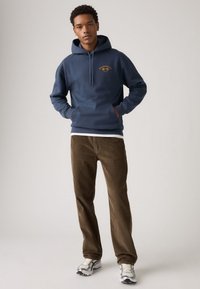 Joven de pie con una sudadera Levi's azul marino, pantalones de pana marrones, camiseta blanca y zapatillas blanco-gris contra un fondo liso.