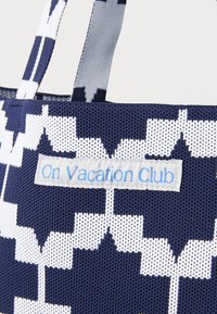 Mornarsko plava i bijela tkanina torba s geometrijskim uzorkom, s svijetloplavom etiketom na kojoj piše "On Vacation Club". Izdržljiv tekstil.