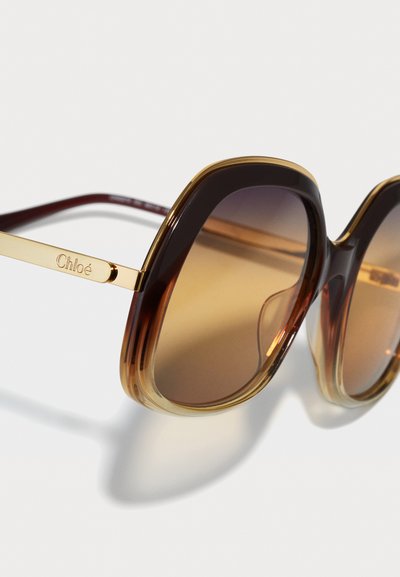 Grandes gafas de sol cuadradas con lentes de color marrón degradado. La montura presenta un diseño transparente en tonos marrón y ámbar con acentos en metal dorado.