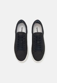Black suede sneakers with flat navy laces, white rubber soles, and a minimalist design. Interior features a contrasting light lining.