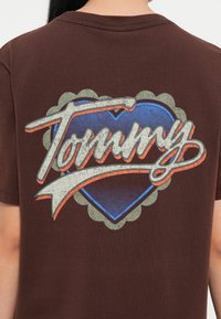 Brown cotton t-shirt featuring a large graphic print of the word "Tommy" in light gray, over a blue heart with orange accents and scalloped border.