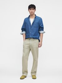 Homme debout portant une chemise en denim bleu sur un t-shirt blanc, un pantalon beige et des mocassins en daim jaune, les mains dans les poches.