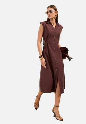 Naine, kes kannab varrukateta pruuni nööpidega midi-kleiti, päikeseprille ja rihmikuid, hoides pruuni käekotti, kõndides ühevärvilise tausta ees.