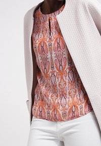 Ljusrosa strukturerad kavaj över en mönstrad ärmlös topp med orange och rosa paisleymönster, tillsammans med vita byxor.