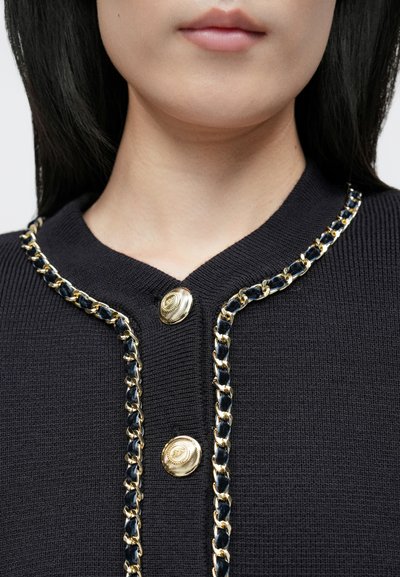 Sötét tengerészkék kardigán bordázott mintával, aranylánc díszítéssel a szélén és nagy, arany gombokkal a nyakkivágásnál.