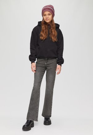 Zwarte hoodie met een ontspannen pasvorm, grijze flared jeans en zwarte platformlaarzen. Het model draagt een kastanjebruine gebreide beanie.