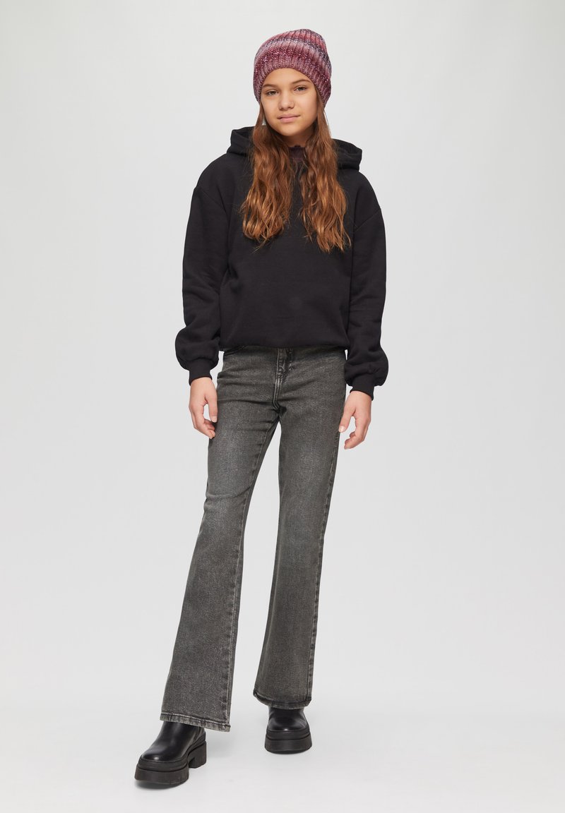 Zwarte hoodie met een ontspannen pasvorm, grijze flared jeans en zwarte platformlaarzen. Het model draagt een kastanjebruine gebreide beanie.