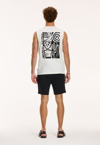 Weißes ärmelloses Shirt mit einem schwarzen Grafi kdesign, das geometrische Formen und Kakteen zeigt. Kombiniert mit schwarzen Shorts und Sandalen.