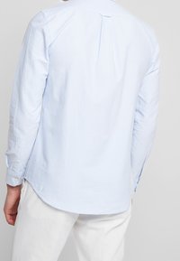 Camisa de manga larga en azul claro, hecha de una tela suave, que presenta un dobladillo redondeado, pliegue en la parte posterior y puños abotonados.