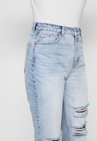 Světlé modré džíny s vysokým pasem, které mají roztrhaný design s dírami a klasický pětivreckový styl.