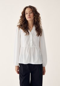 Woman with curly hair wearing a white long-sleeve blouse with ruffle trim and black pants, standing against a plain light background.