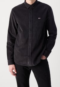 Camisa de botón de pana negra con cuello, que presenta un pequeño logo en el lado izquierdo del pecho y mangas largas. Tejido texturizado con líneas de relieve.
