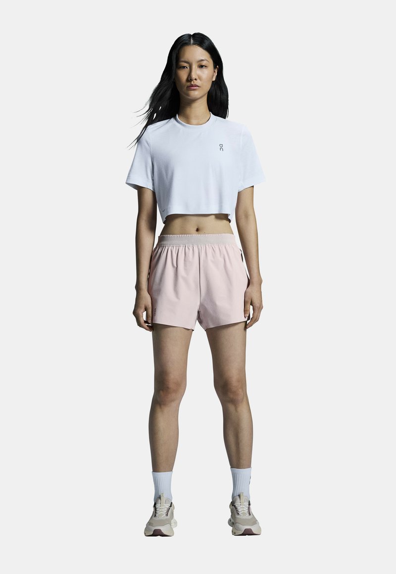 Weißes, kurz geschnittenes T-Shirt mit Logo, kombiniert mit hellpinkfarbenen Sportshorts mit elastischem Bund. Weiße Socken und neutrale Sneakers runden das Outfit ab.