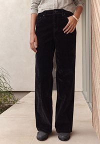 Pantalones de pana negros con un diseño de pierna recta, que cuentan con bolsillos delanteros, un ligero brillo y una cintura alta. Usados con calzado gris.