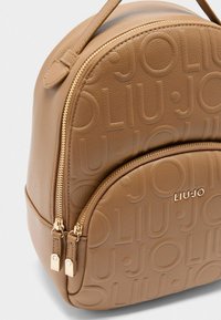 Zaino in ecopelle color tan con motivo logo in rilievo, forma arrotondata, manico superiore e due scomparti con cerniera in metallo dorato.