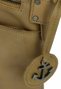 Bolso de cuero marrón claro con un bolsillo con cremallera, acentuado con herrajes de plata y un charm en forma de ancla circular adjunto a una correa. Textura suave.