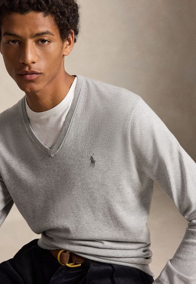 Jonge man die een lichtgrijze V-hals trui met een klein logo draagt, een wit ondershirt, donkere broek en een bruine riem tegen een neutrale achtergrond.