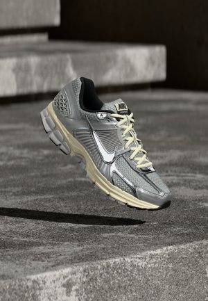 Szürke és bézs Nike futócipő fehér fűzőkkel és nagy fehér Nike logóval, mintha egy betonfelület fölött lebegne.
