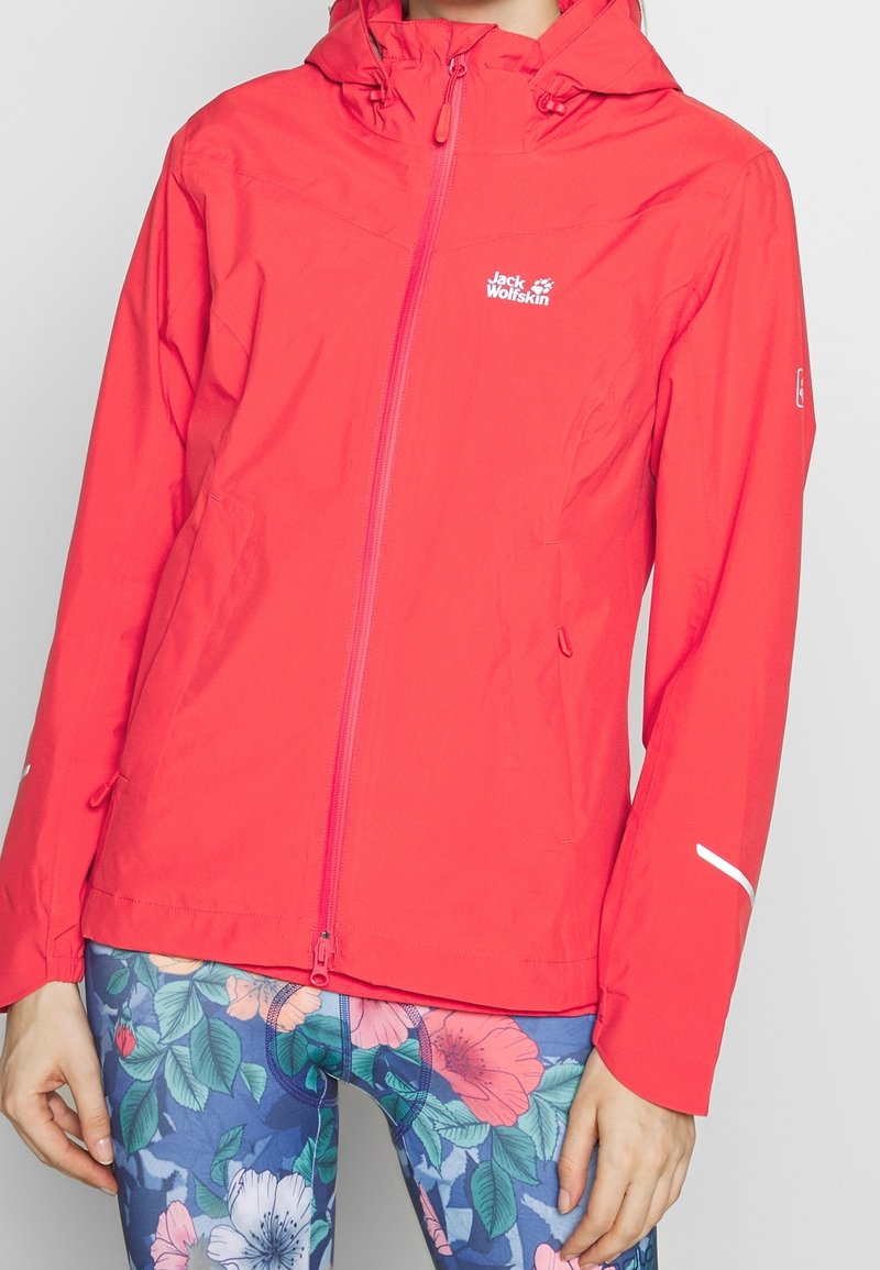 Giacca impermeabile rossa con zip frontale e cappuccio regolabile. Presenta dettagli riflettenti e un logo sul petto. Abbinata a leggins floreali.